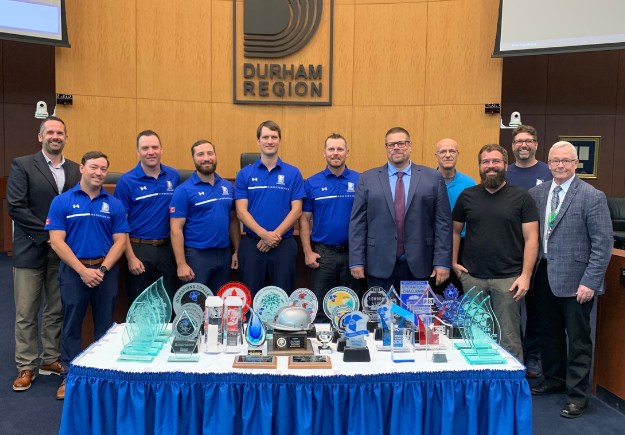 A photo of Durham Region’s Sludgehammer team in blue shirts standing behind a table of trophies at a Council Meeting on September 24, 2025.