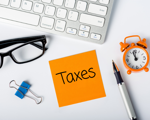 Flat lay image of keyboard, glasses, paper clip, pen, clock, and sticky note with writing on it that reads “Taxes”.