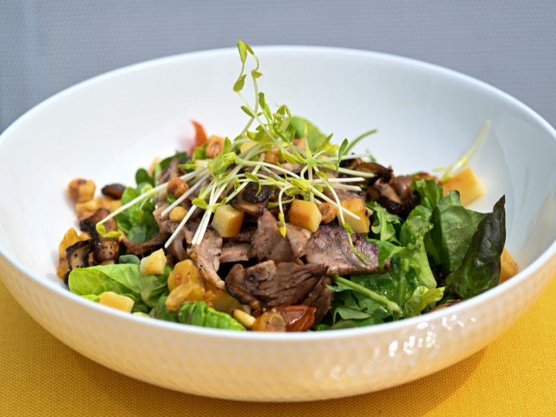 Image of a harvest bowl with fresh greens and roasted vegetables, topped with grilled steak and micro greens