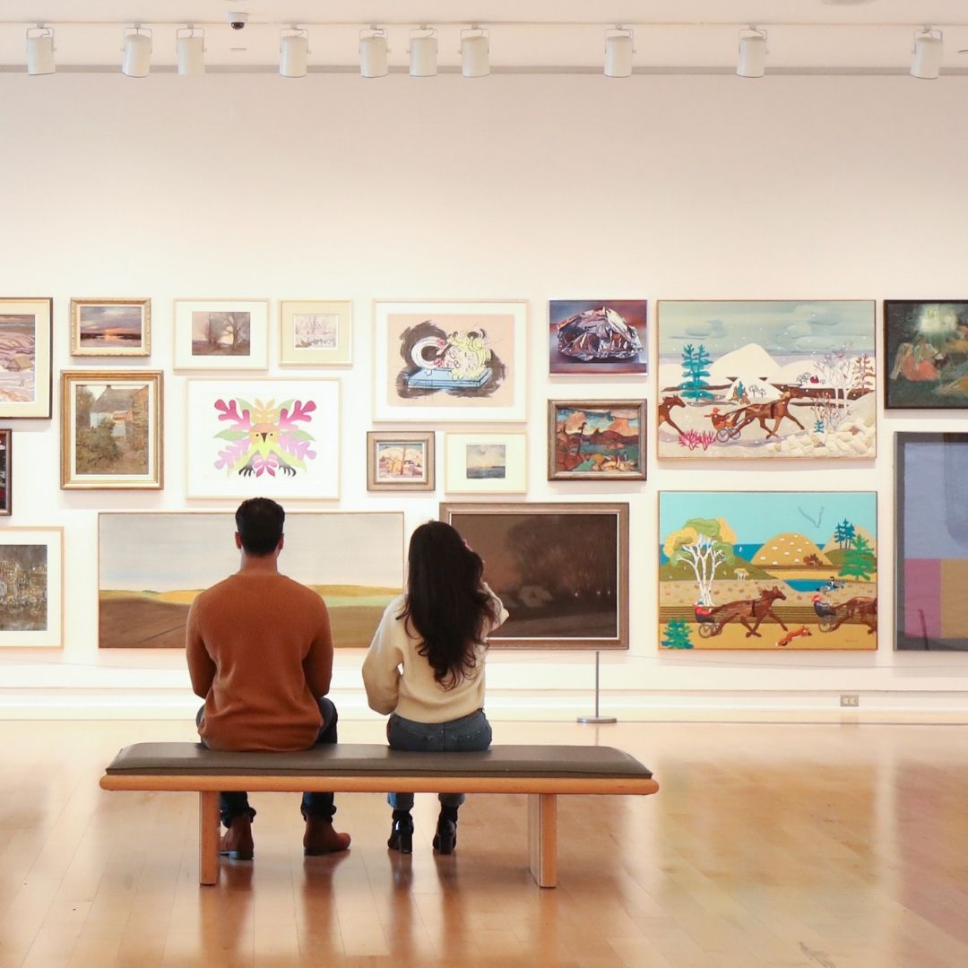 Image of two people sitting on a bench at an art gallery taking in a wall full of framed paintings