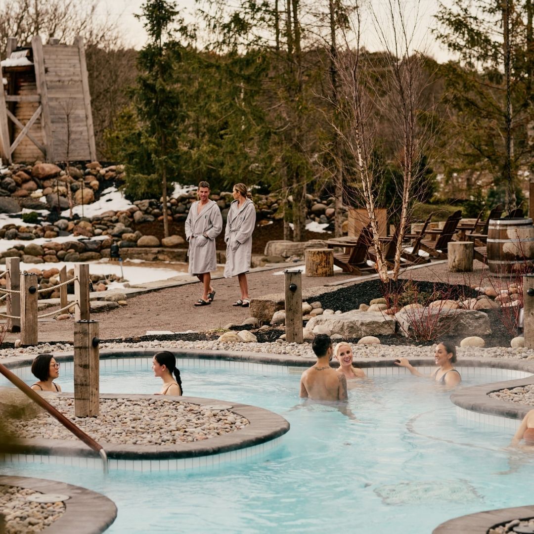 Image of an outdoor Nordic spa with patrons in bath robes and blue steamy hot tubs