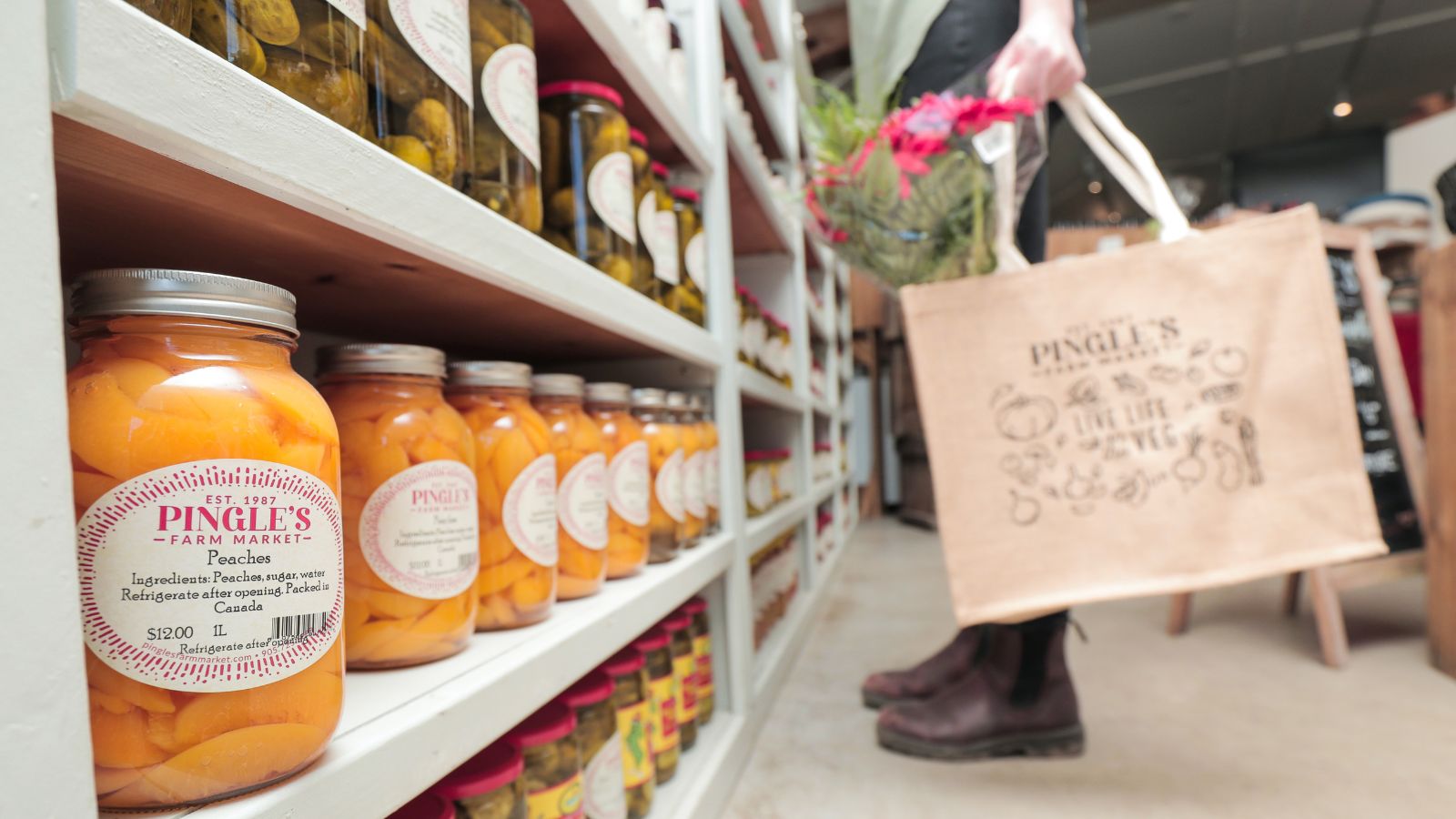 Image of a person from the knees down standing in front of a wall of jarred preserves and pickles holding a tote bag