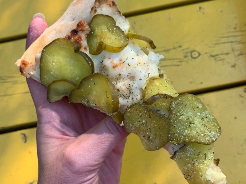 Image of a person holding a slice of pizza topped with sliced pickles