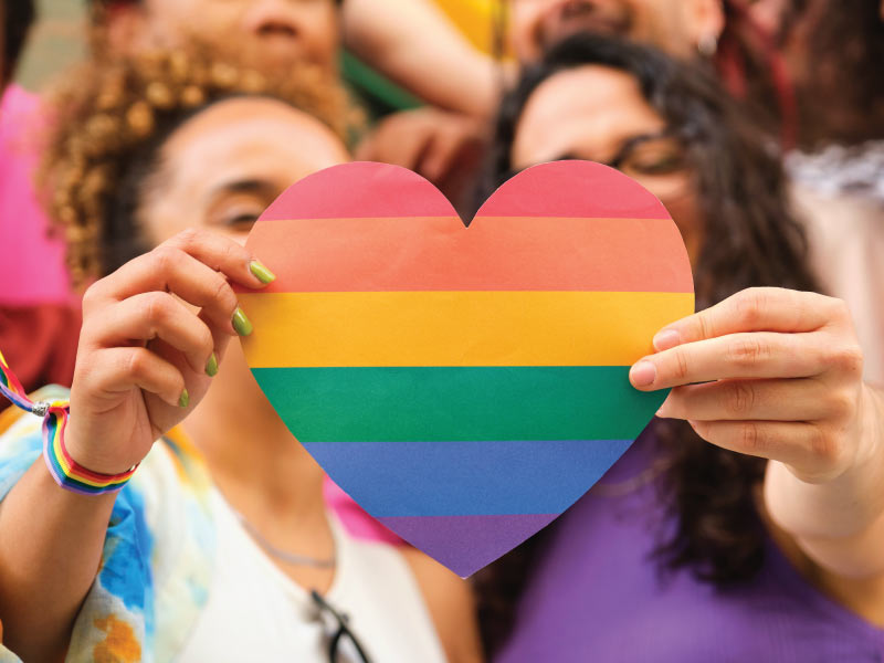 A couple holding a paper rainbow heart in front of their faces.