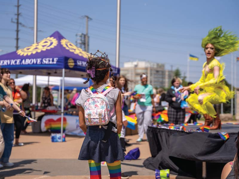 A young girl with a backpack and rainbow accessories watching a performance on a stage outside.